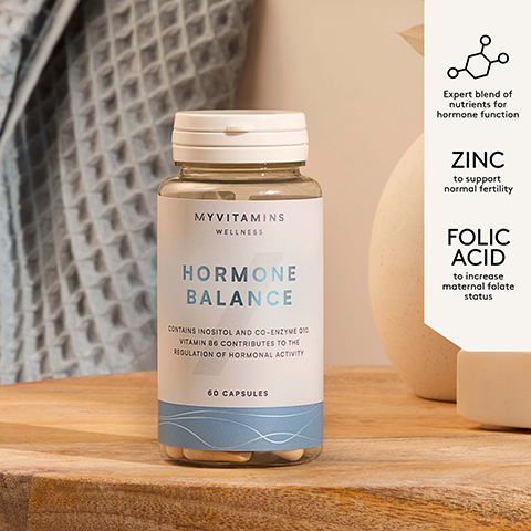 A bottle of MYVITAMINS WELLNESS HORMONE BALANCE 60 CAPSULES, which states CONTAINS INOSITOL AND CO-ENZYME Q10. VITAMIN B6 CONTRIBUTES TO THE REGULATION OF HORMONAL ACTIVITY, is displayed on a wooden surface with a blurred background of a light towel and cream jug, accompanied by a white information panel detailing Expert blend of nutrients for hormone function, ZINC to support normal fertility, and FOLIC ACID to increase maternal folate status.