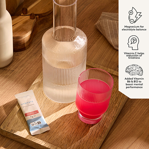 A Myvitamins Hydrate sachet, a ribbed glass with pink liquid, and a ribbed carafe of water are arranged on a wooden board, with a side panel displaying product benefits; the sachet reads Myvitamins Wellness Hydrate With Electrolytes Strawberry & Cherry Flavour 7g e and FR boite+sachet, while the panel states Magnesium for electrolyte balance, Vitamin C helps reduction of tiredness, and Added Vitamin B6 & B12 to boost mental performance.