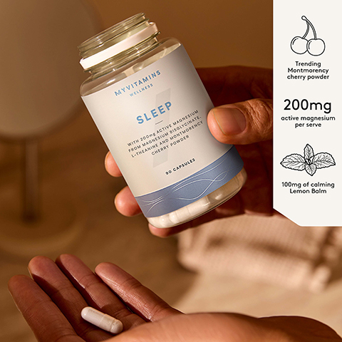 A hand holds a clear bottle of Myvitamins supplements, tilted to dispense a white capsule into another cupped hand below, with visible text reading Myvitamins Wellness SLEEP WITH 200mg ACTIVE MAGNESIUM FROM MAGNESIUM BISGLYCINATE. L-THEANINE AND MONTMORENCY CHERRY POWDER 90 CAPSULES Trending Montmorency cherry powder 200mg active magnesium per serve 100mg of calming Lemon Balm.
